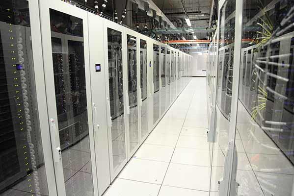 Modern data center with rows of tall server racks lined up in a brightly lit room. The servers are enclosed in white cabinets with glass doors, and small indicator lights are visible on the equipment. The floor is made of white tiles, and the ceiling is equipped with various cables and equipment for cooling and power.