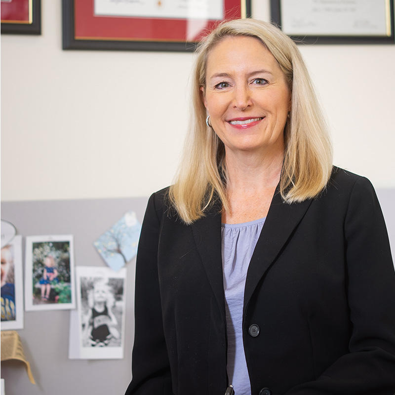 A professional headshot of Dr. Lynne Wagner, a White woman with a light skin tone and blonde hair, wearing a black blazer and blue top while smiling at the camera and standing in her office.