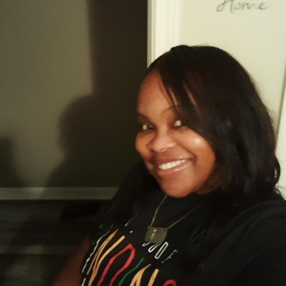 A black woman wearing a black t-shirt and chain necklace, taking a selfie in her home.
