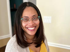 A woman, Dr. Robin Lockridge, with glasses and straight brown hair wearing a yellow shirt and gray cardigan sits at her laptop smiling at the camera.