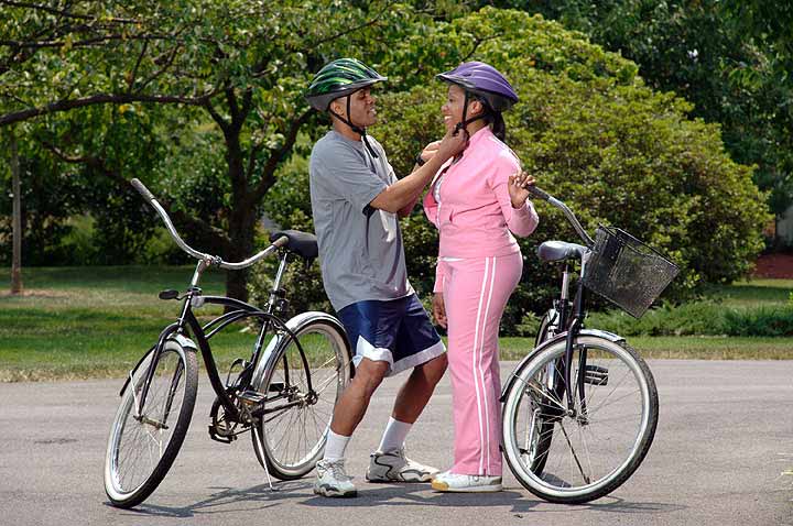 An African-American man adjusts an African-American woman's helmet strap as they prepare for a bicycle ride.