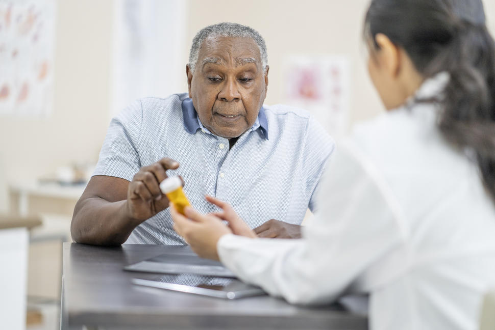 En la foto se observa a un adulto mayor sentado frente a un profesional médico que sostiene un frasco de medicamento de venta con receta. Ambos parecen estar conversando sobre el medicamento.