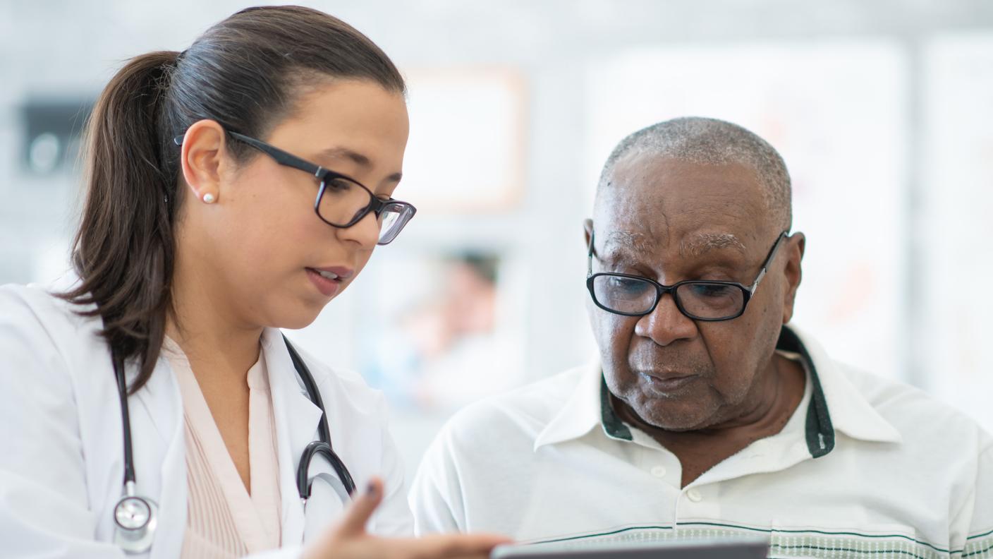 Una joven doctora muestra información en una tableta a un paciente negro de edad más avanzada.